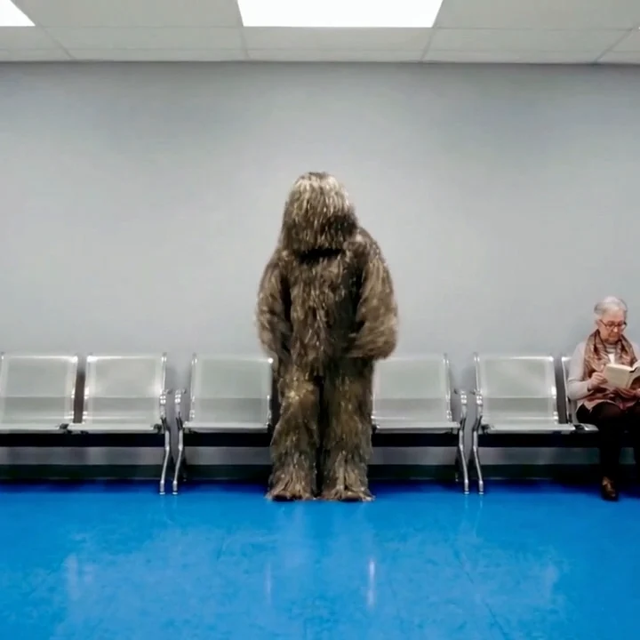 A figure in a ghillie suit standing in a blue-floored waiting room.